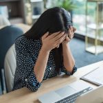 Mulher branca setada em uma mesa, em frente ao computador, triste/esgotada