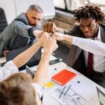 5 pessoas sentadas em uma mesa de ambiente corporativo. Todas com o braço levantado, em direção ao meio.