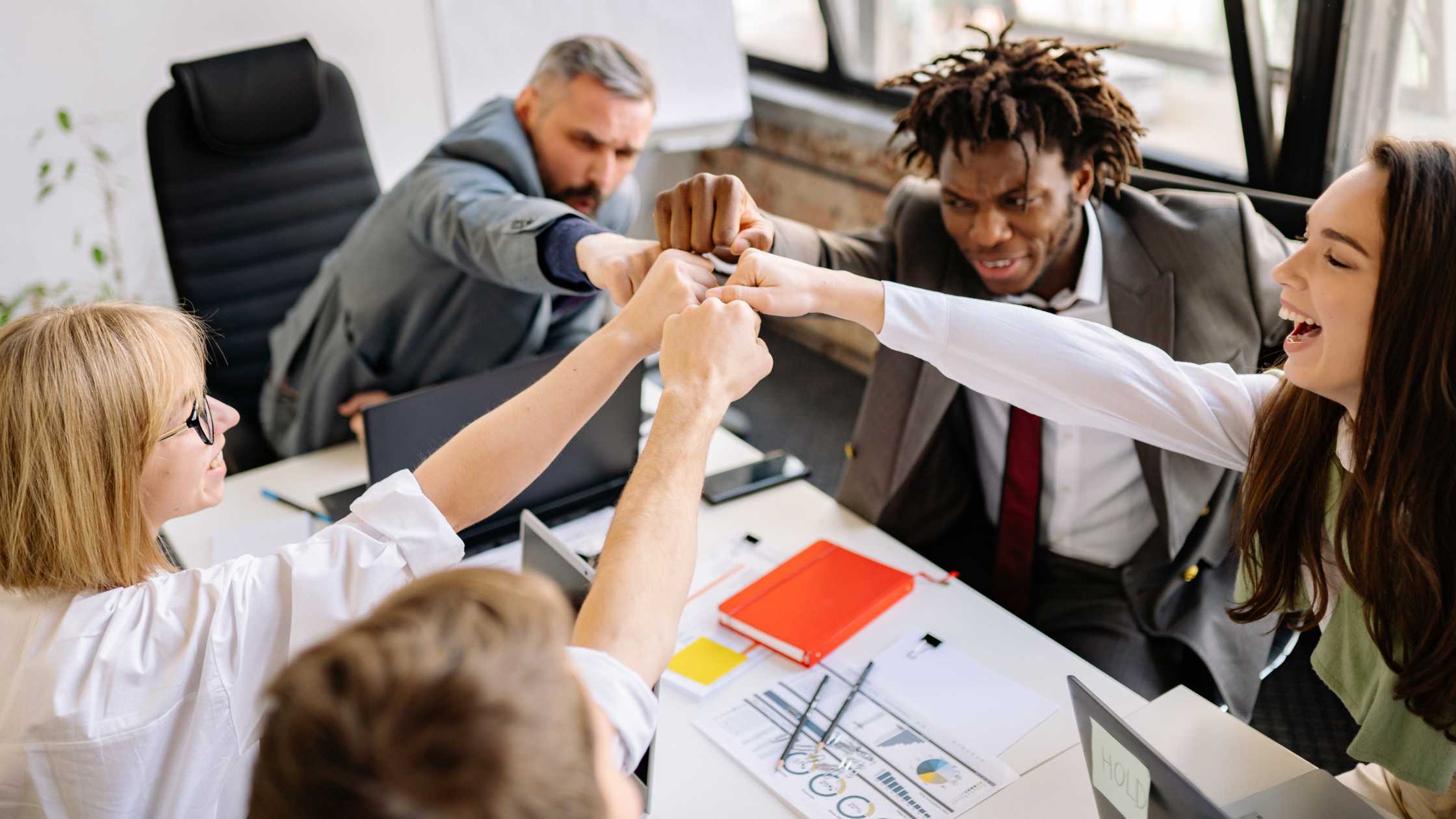 5 pessoas sentadas em uma mesa de ambiente corporativo. Todas com o braço levantado, em direção ao meio.