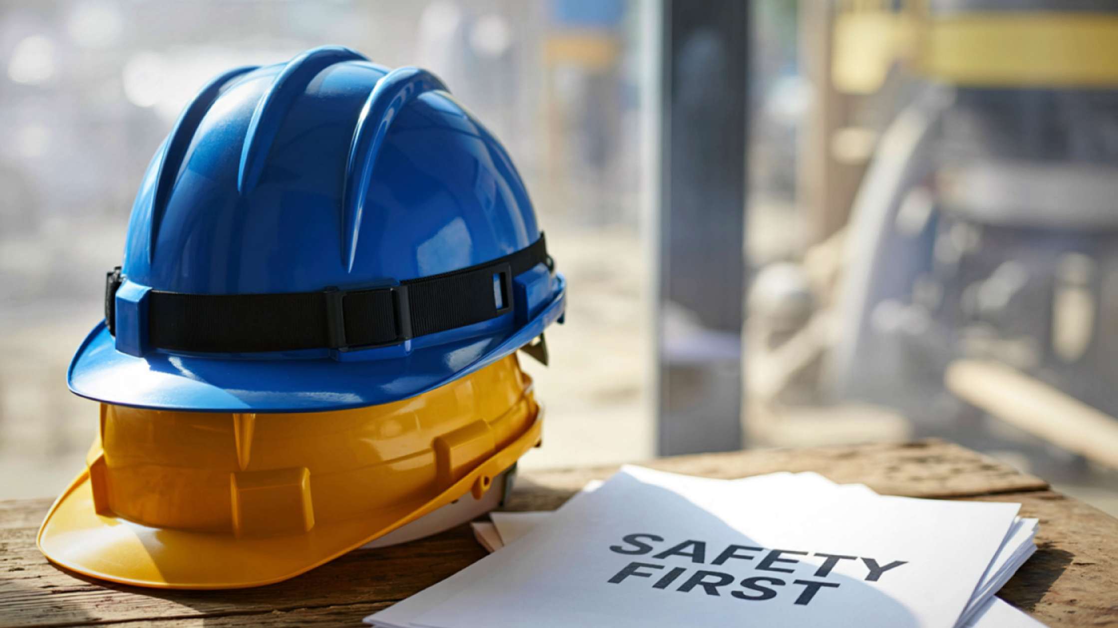 Dois capacetes de segurança empilhados, em cima de uma mesa, com uma folha escrito "Segurança primeiro". Fundo de uma obra.