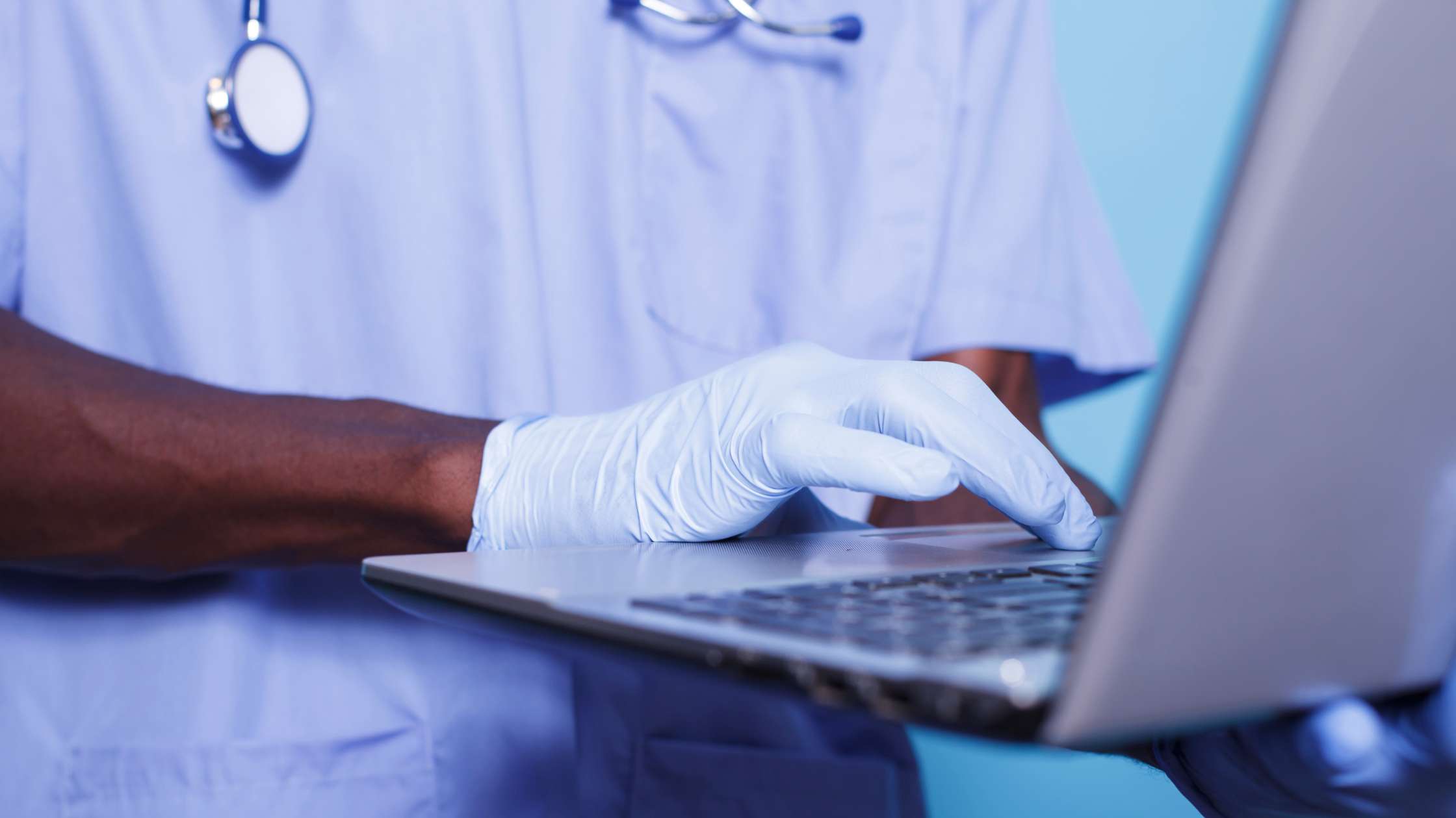 Pessoa negra com um computador na mão, e equipada com luvas descartáveis, jaleco e estetoscópio no pescoço.